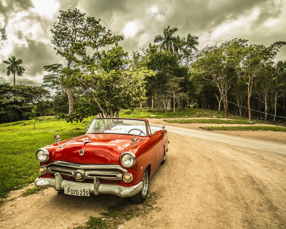 Bäume, bewölkter Himmel und ein roter klassischer Oldtimer auf nicht asphaltierter Straße